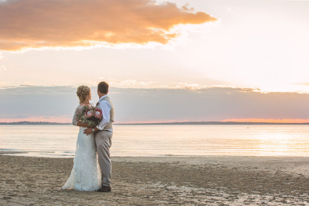 bride and groom watch colourful cloudy sunset by beach