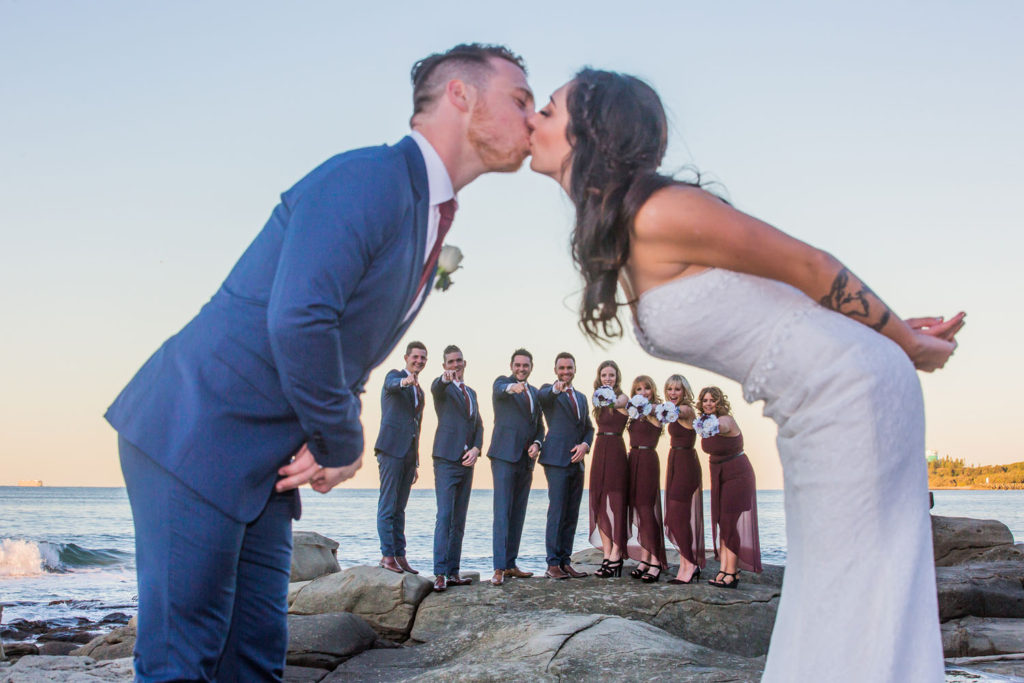 photograph of bride and groom kissing with wedding party at beach