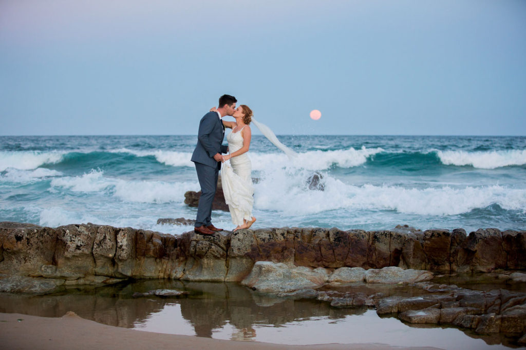 photograph of wedding couple kissing by ocean with moon